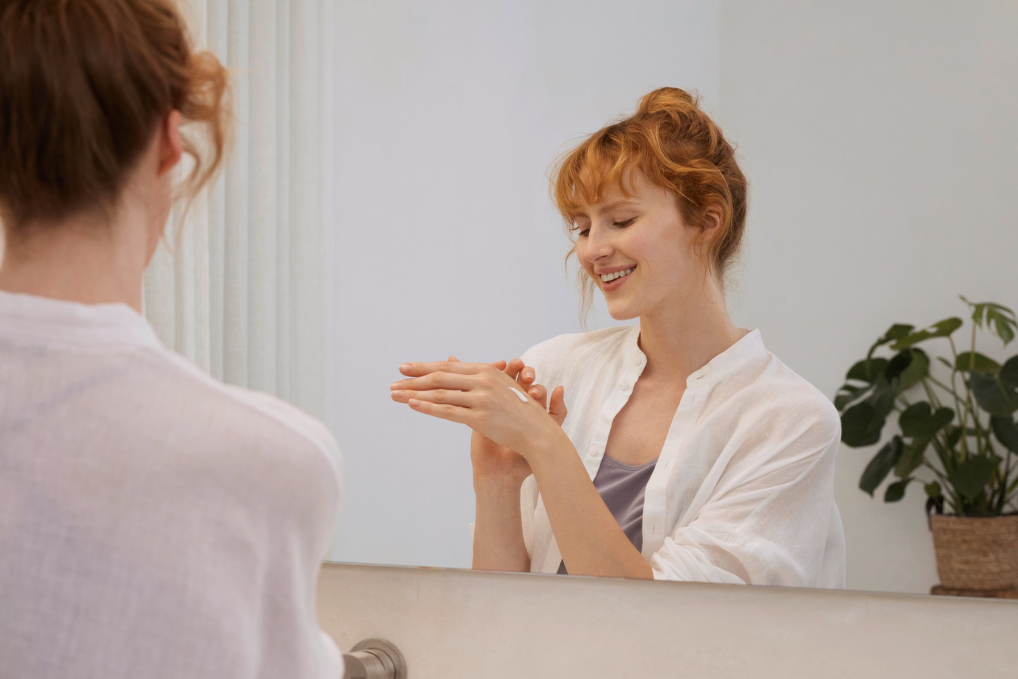 Femme aux cheveux roux se crème les mains en souriant devant le miroir