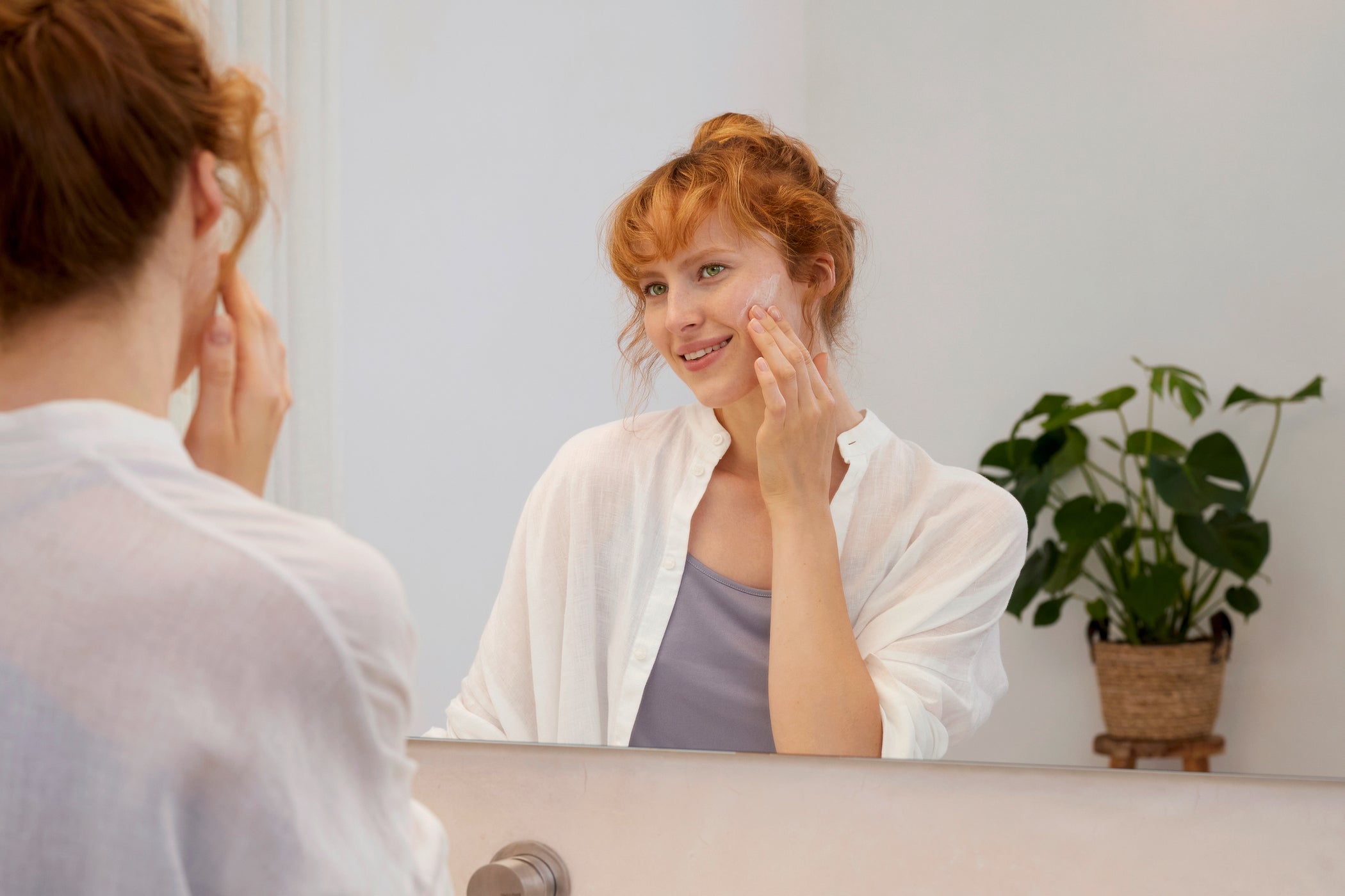 Femme rousse debout dans la salle de bain devant le miroir appliquant de la crème sur la joue