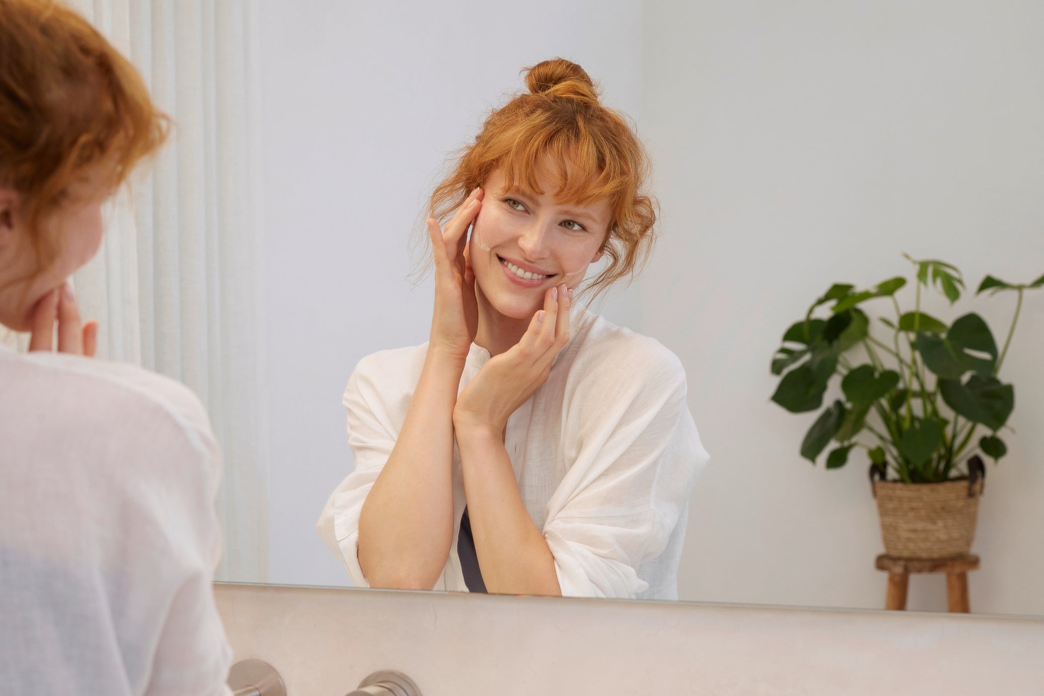 Jeune femme aux cheveux rougeâtres dans la salle de bain devant le miroir et s'applique de la crème sur le visage avec les deux mains