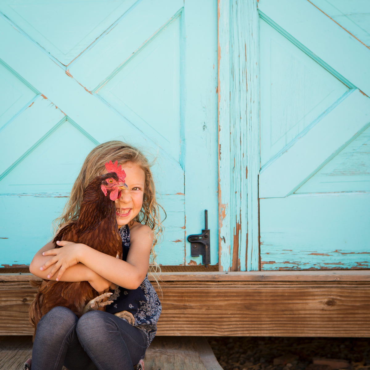 Enfant enlace une poule brune devant une porte en bois turquoise, souriant