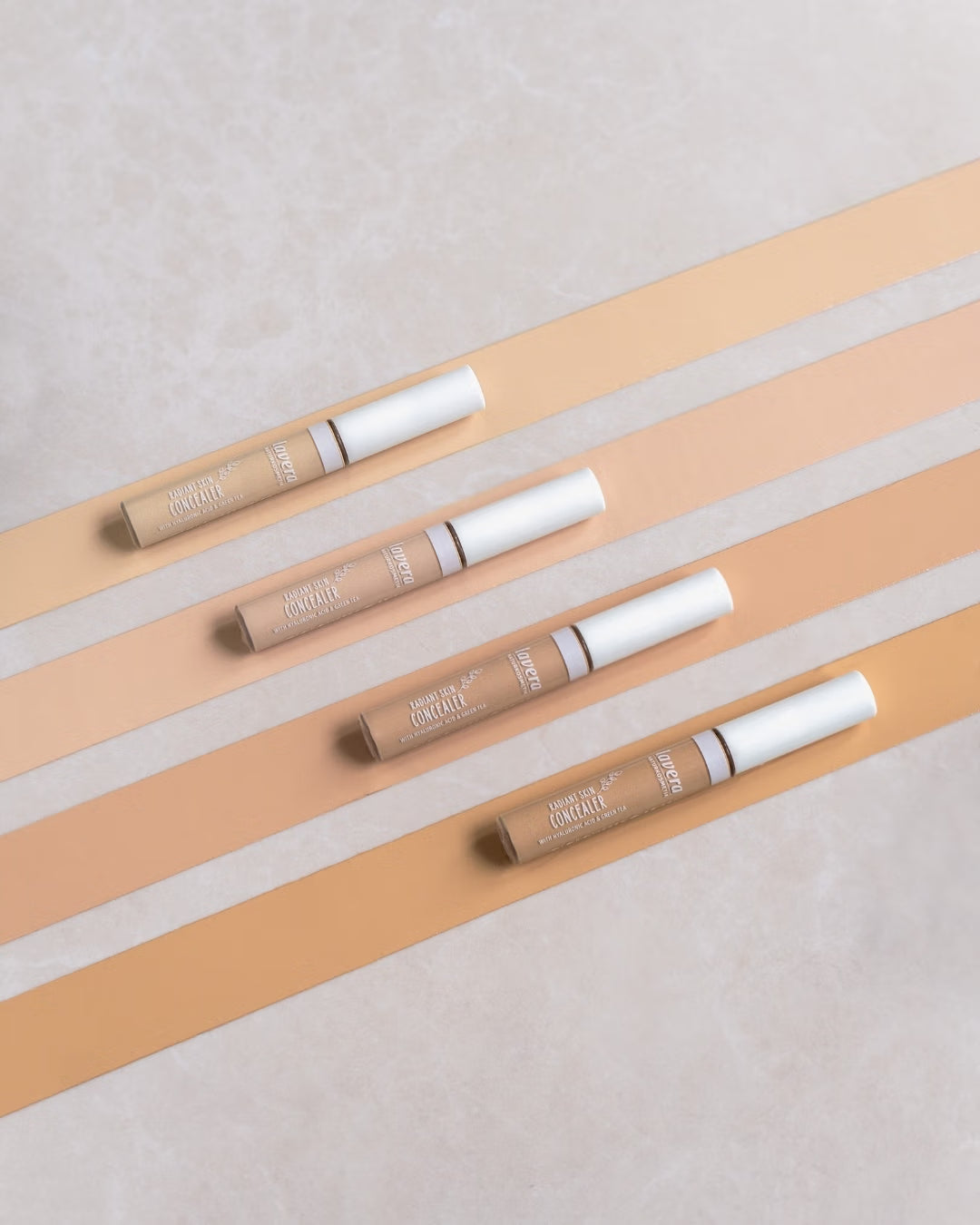 Vier lavera Radiant Skin Concealer in verschiedenen Farbtönen auf einem beigefarbenen Streifenmuster.
