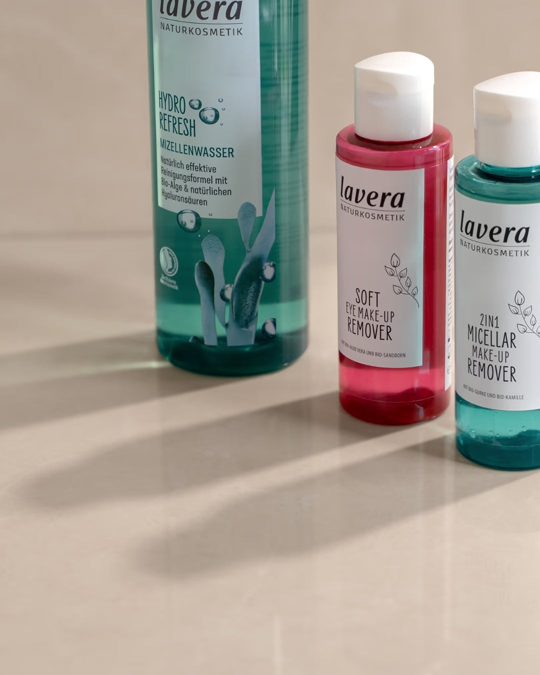Drei lavera Reinigungsprodukte – Hydro Refresh Mizellenwasser sowie Soft und 2in1 Mizellen-Make-up-Remover – stehen auf einer beige schimmernden Oberfläche.