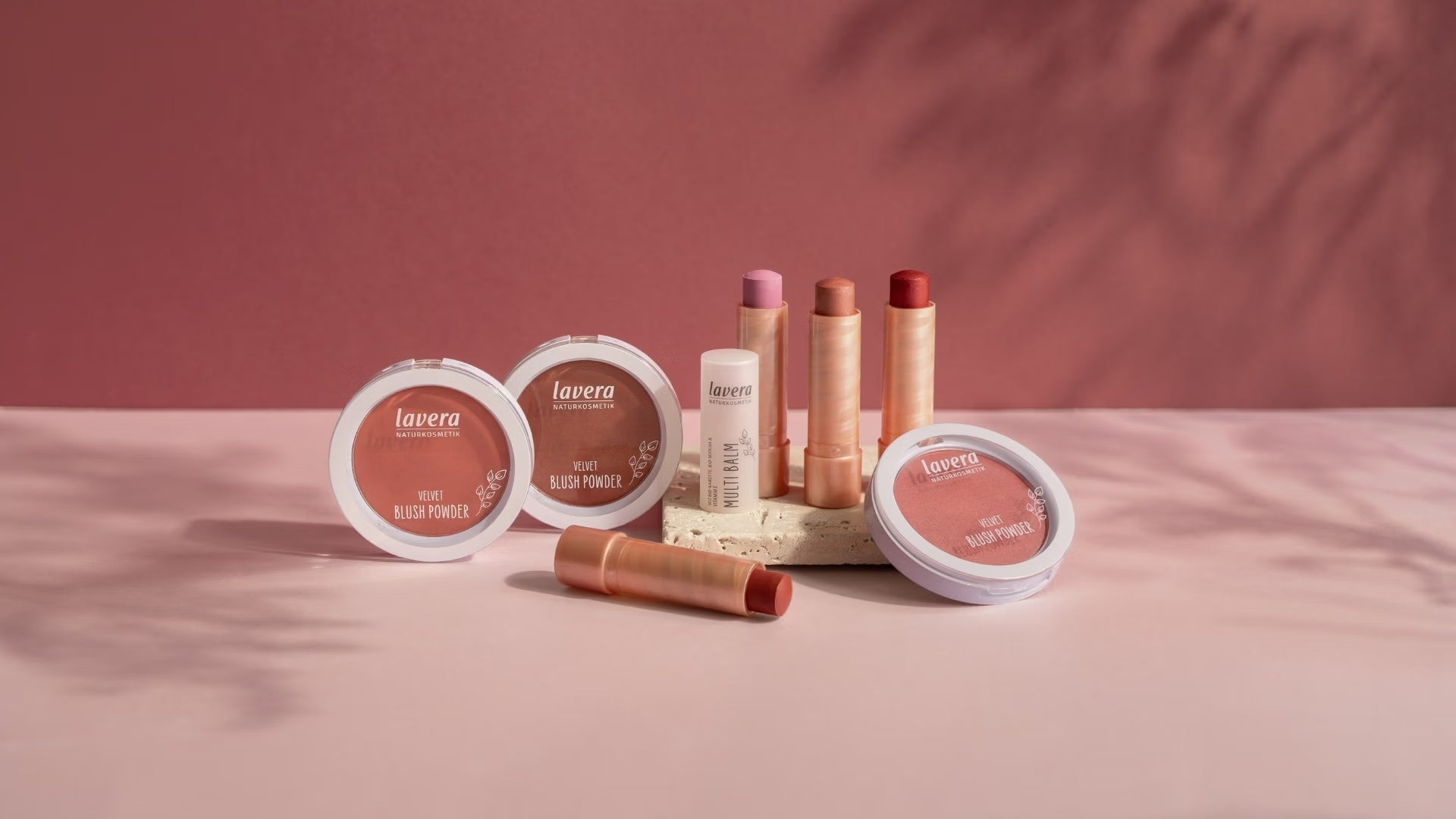 Mehrere lavera Rouge-Produkte und Multi Balms in Rosé-, Nude- und Rottönen stehen auf einem Podest vor rosafarbenem Hintergrund.
