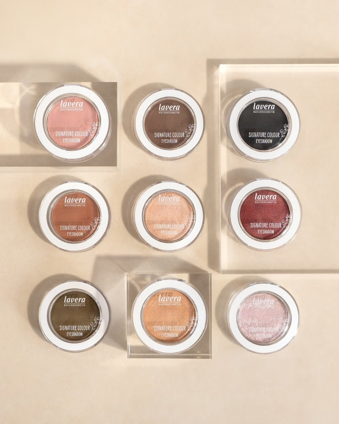 Neun lavera Signature Colour Lidschatten in verschiedenen Farbtönen liegen auf transparenten Acrylblöcken vor beigem Hintergrund.