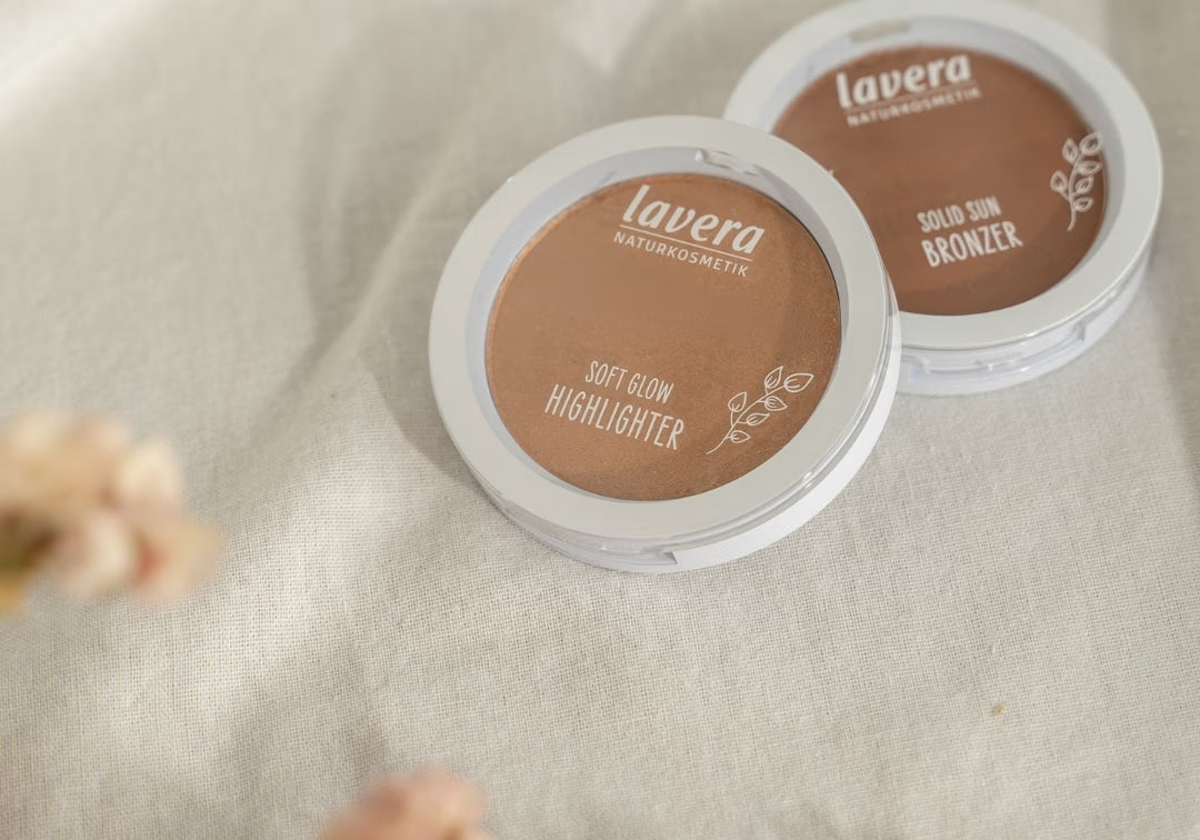 lavera Teintprodukte, ein Soft Glow Highlighter und ein Solid Sun Bronzer, liegen auf hellem Leinenstoff neben unscharfen rosafarbenen Blüten.