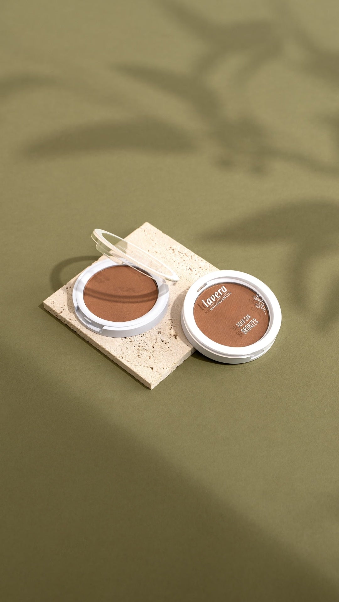 Zwei geöffnete lavera Teintprodukte – Highlighter und Solid Sun Bronzer – liegen auf einem hellen Steinpodest vor olivgrünem Hintergrund.