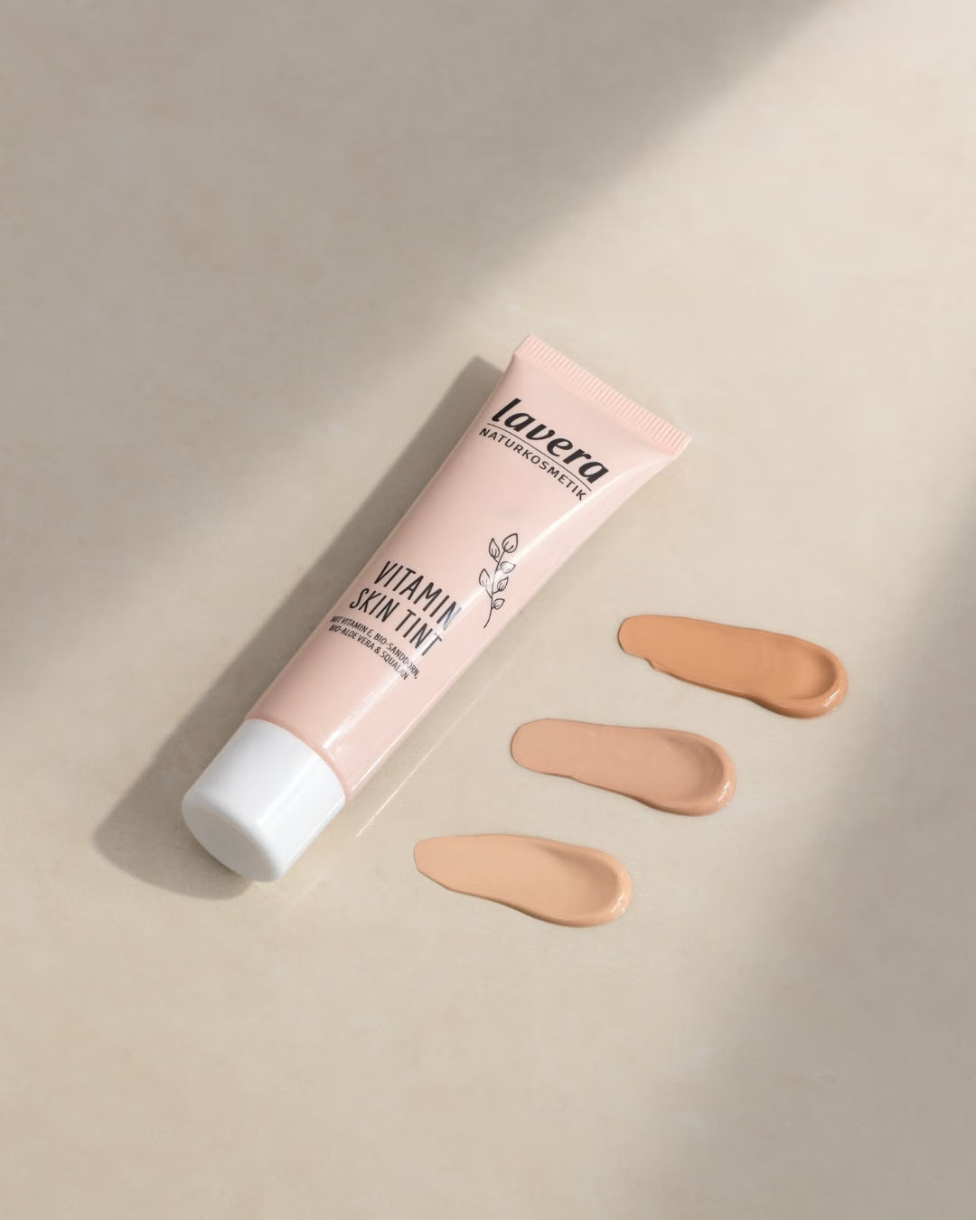 Eine Tube lavera Vitamin Skin Tint liegt neben drei unterschiedlich hellen Farbtupfern auf einer beigefarbenen Oberfläche.