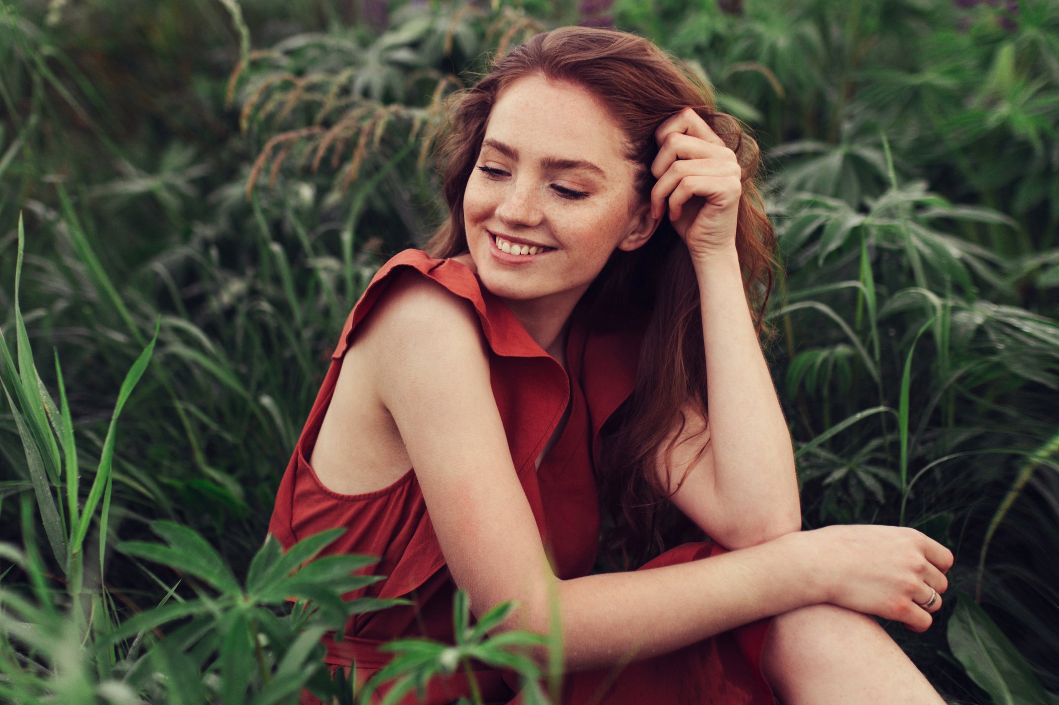 Femme rousse en robe rouge assise dans l'herbe et souriante, portrait nature