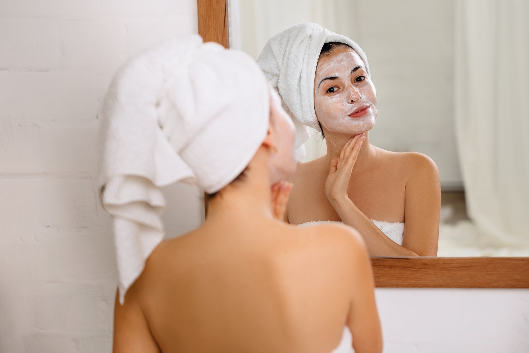 Femme avec serviette dans les cheveux applique masque facial blanc, regard dans le miroir
