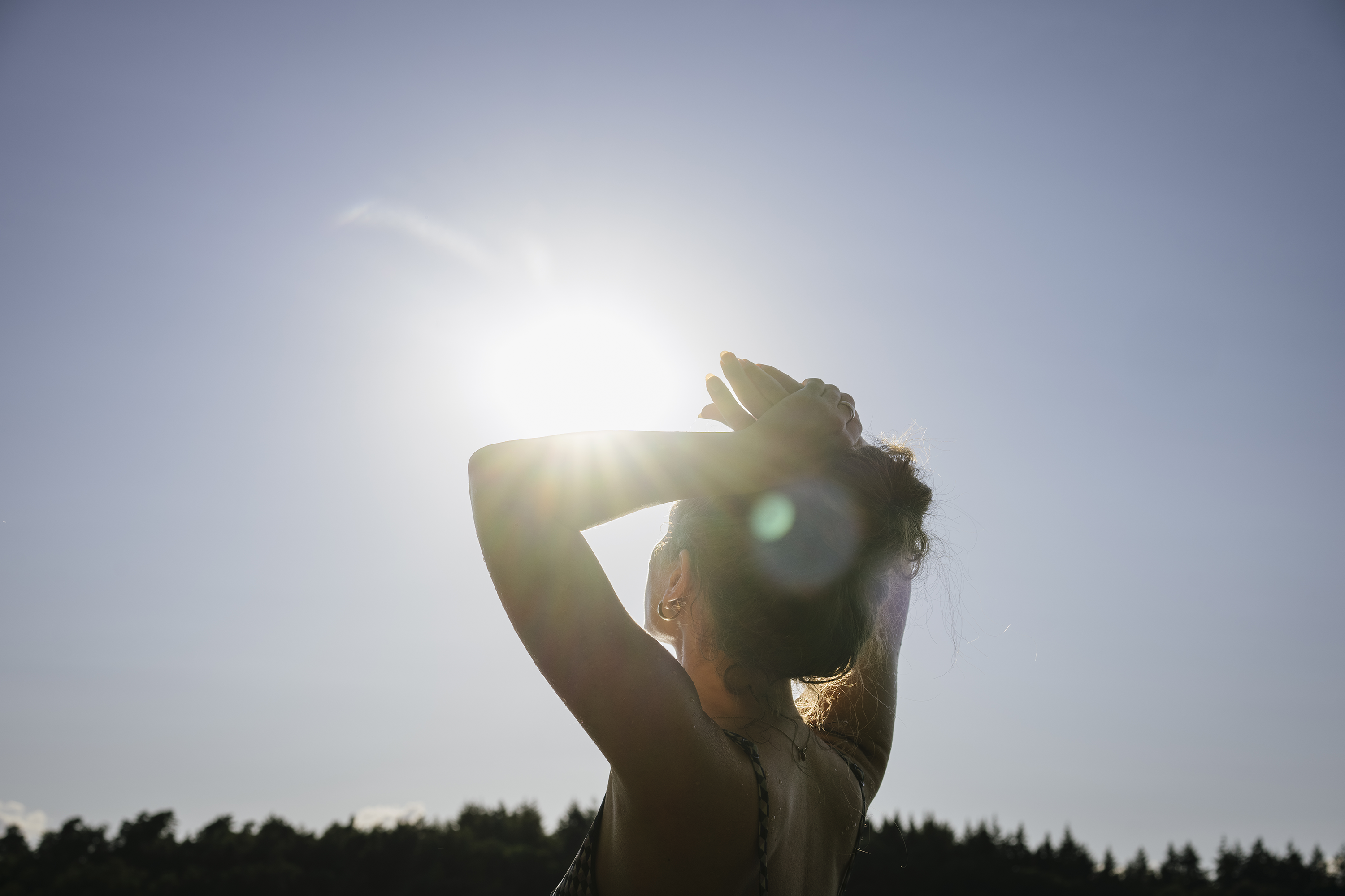 Femme debout à contre-jour du soleil, tient ses cheveux en hauteur et regarde le ciel lumineux