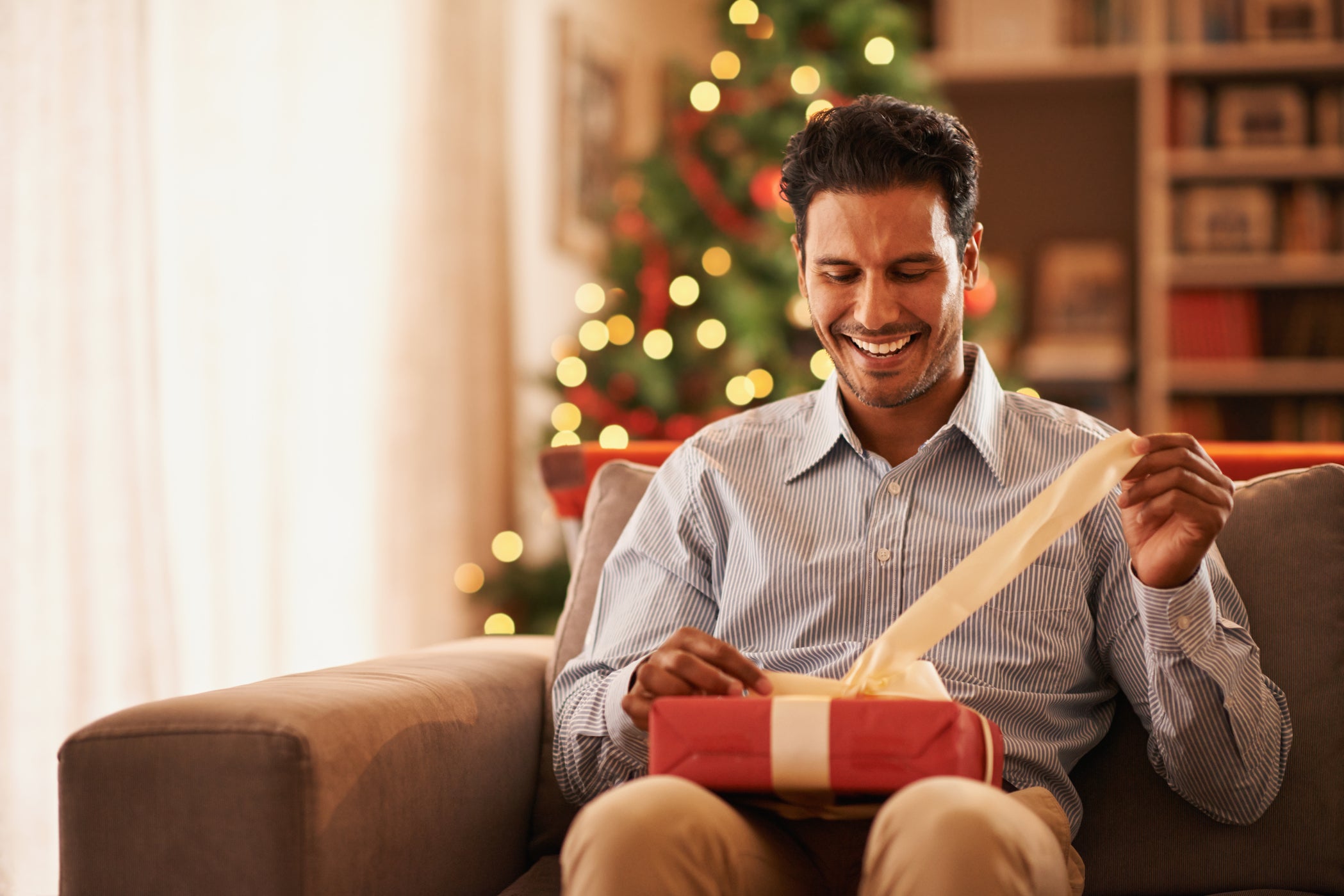 Homme assis sur un canapé déballant un cadeau – avec sapin de Noël en arrière-plan