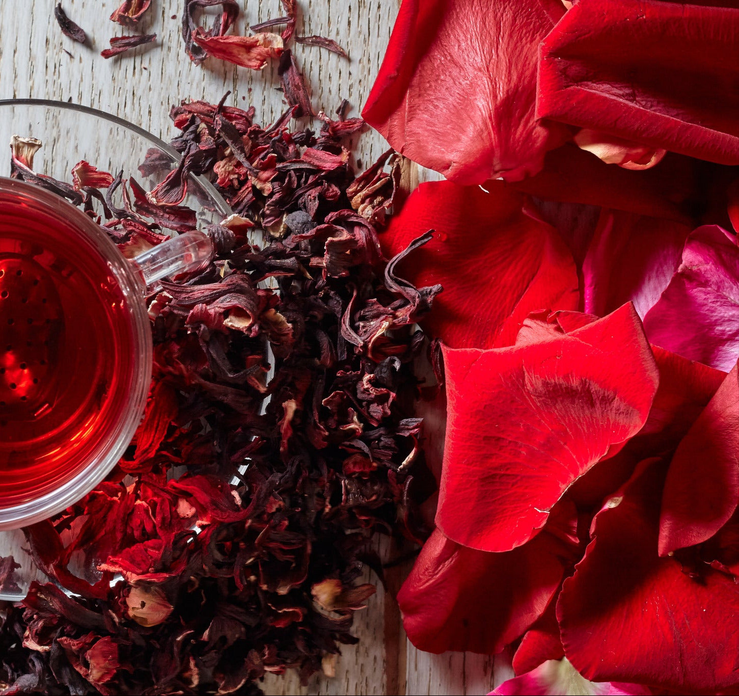Frisch aufgebrühter Hibiskus-Tee in einer Glastasse mit Herz-Teesieb, umgeben von getrockneten Hibiskusblüten und roten Blütenblättern.